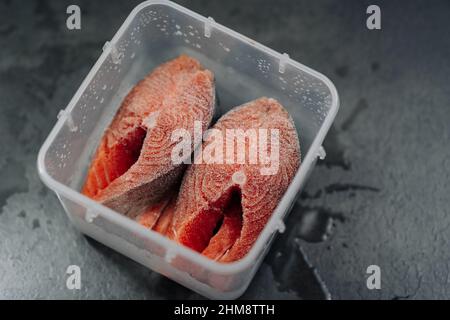 Blick von oben auf frisch gefrorenen rohen roten Lachsfisch. Gefrorene Lachssteaks in einer Plastikbox oder einem Behälter auf dunkelgrauem Hintergrund. Stockfoto