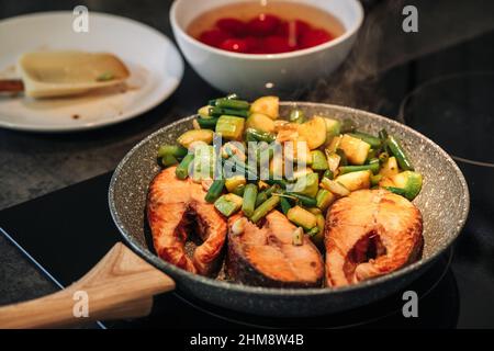 Nahaufnahme einer unkenntlichen Frau, die das Mittagessen in der Küche zubereitet. Gegrilltes Forellensteak. Braten unter Rühren. Die Zucchini und die grünen Bohnen werden in der Pfanne gebraten. Strom Stockfoto