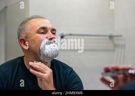 Gut aussehender reifer Mann mit grauen Haaren versucht, seinen Bart selbst in einem Badezimmer abzuschneiden Stockfoto