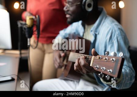 Nahaufnahme eines afrikanischen Mannes in Kopfhörern, der Gitarre spielt, während er gegenüber dem Mikrofon sitzt und eine Frau im Hintergrund steht Stockfoto