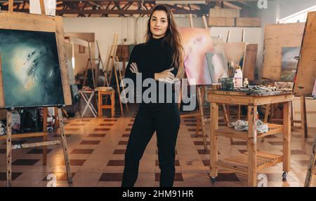 Professionelle Malerin, die in ihrer Werkstatt steht und ihre Kunstwerke hinter sich hat. Selbstbewusste Künstlerin, die mit ihren Armen auf die Kamera schaut Stockfoto