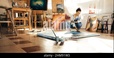 Künstler macht ein blaues Gemälde auf einer Leinwand. Die Malerin berührt ihr Gemälde, während sie in ihrem Atelier auf dem Boden hockt. Kreative junge Frau w Stockfoto