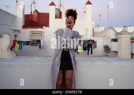Fröhliche afroamerikanische Frau in trendigem Casual-Outfit mit lockigem Haar lachend über Witz, während sie sich abends auf der Terrasse des Wohnhauses ausruhte Stockfoto
