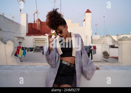 Afroamerikanische Frau in stilvoller Kleidung, die Sonnenbrille anpasst und sich an die Schranken an Häusern lehnt und am Abend den wolkenlosen Sonnenuntergang in der Sonne von caisz, Spa, genießt Stockfoto