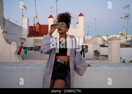 Afroamerikanische Frau in stilvoller Kleidung, die Sonnenbrillen anpasst und sich an der Barriere gegen die Häuser lehnt, während sie sich abends auf der Terrasse in cadez, Sp Stockfoto