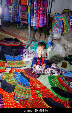 Ein süßer, einheimischer Junge sitzt in einem Verkaufsstand, der Stoffe und Souvenirs auf dem Nachtmarkt in der Fußgängerzone im Zentrum von Luang Prabang, Laos, Südostasien, verkauft Stockfoto