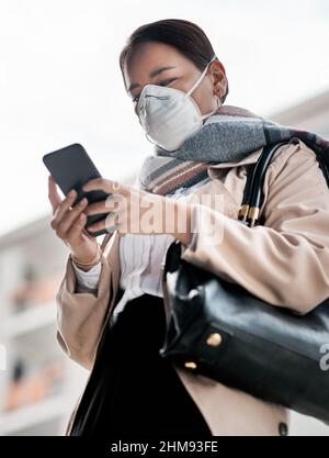 Diese App zur Verfolgung macht mich beim Ausgehen noch mehr wohl fühlen. Aufnahme einer jungen Frau, die eine Maske trägt und ein Smartphone benutzt, während sie in der Stadt unterwegs ist. Stockfoto