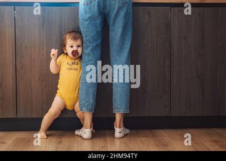 Porträt eines kleinen Mädchens, das die Beine der Mutter zu Hause hält. Babypflege. Stockfoto