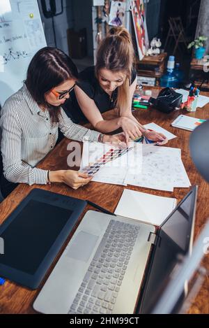 Junge Grafikdesignerinnen sitzen mit Laptop, Farbkarten und einigen Skizzen am Schreibtisch und diskutieren über das neue Projekt, das ein Konzept auswählt. Stockfoto