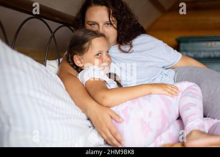 Junge kaukasische Tochter und Mutter, die sich umarmen und auf einem Doppelbett liegen, blicken beide in einem Holzhaus mit Pyjamas tagsüber auf die Kamera. Familie Stockfoto