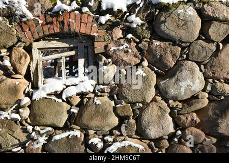 Ein Fragment einer alten, halb zerstörten, schneebedeckten Steinmauer mit einem weißen Holzfenster an einem sonnigen Wintertag in Europa. Hintergrundbild Stockfoto
