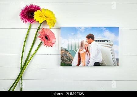 Leinwanddruck mit Galerie Wrap Technik auf weißem Holztisch, Draufsicht. Hochzeitsfoto und Blumenstrauß von Gerbera Blumen. Gestreckte Fotografie. Portrai Stockfoto