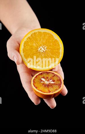 Halbierte orangefarbene Früchte in den Händen auf schwarzem Hintergrund. Person, die zwei Hälften verschiedener Orangen hält. Reife Zitrusfrüchte mit gelbem und rotem Juic Stockfoto
