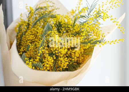 Stilvolles Mimosa Bouquet auf weißem Hintergrund. Glücklicher Frauentag und glücklicher Muttertag. Wunderschöne Mimosenblumen in Bastelpapier in der Nähe des Fensters. 8 ma Stockfoto