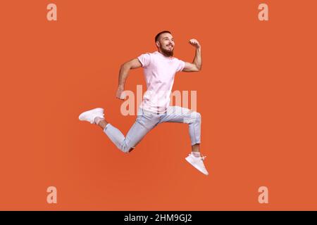 Ganzkörperportrait mit positiv inspiriertem Bartmann, der in der Luft springt oder schnell läuft und Glück zum Ausdruck bringt, in einem rosa T-Shirt. Innenaufnahme des Studios isoliert auf orangefarbenem Hintergrund. Stockfoto