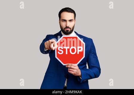 Ernsthafter bärtiger Mann, der auf die Kamera zeigt und das Schild Stop Road als Symbol für Verbot, Einschränkungen hält und einen offiziellen Anzug trägt. Innenaufnahme des Studios isoliert auf grauem Hintergrund. Stockfoto