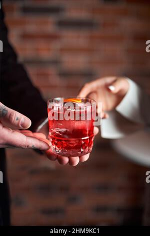 Red Negroni Cocktail mit orangefarbener Schale in einem altmodischen Glas, in der Hand eines Mannes, der es einer Freundin anbietet Stockfoto