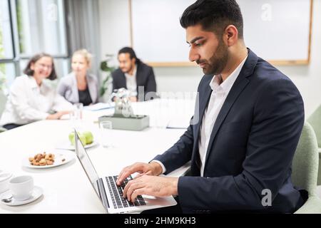 Konzentrierter Geschäftsmann, der im Konferenzraum mit Team im Hintergrund auf einem Laptop schreibt Stockfoto