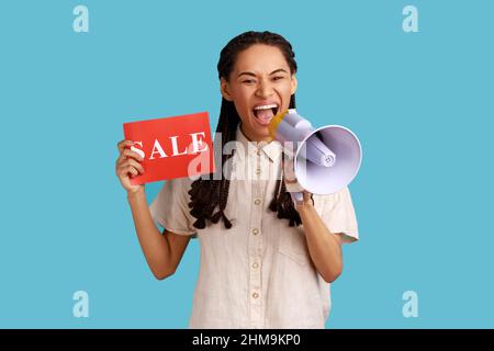 Porträt einer emotionalen Frau mit schwarzen Dreadlocks, die eine Karte mit Verkaufsaufschrift hält und im Megaphon über Rabatte schreit, in weißem Hemd. Innenaufnahme des Studios isoliert auf blauem Hintergrund. Stockfoto