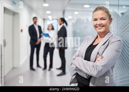 Erfolgreiche Geschäftsfrau, die mit gekreuzten Armen vor dem Geschäftsteam im Büro steht Stockfoto