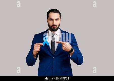 Bewusstsein für Prostatakrebs. Ernsthafter Mann, der in seinen Händen auf das blaue Band zeigt, die Menschen unterstützt, die leben und krank sind, trägt einen Anzug im offiziellen Stil. Innenaufnahme des Studios isoliert auf grauem Hintergrund. Stockfoto