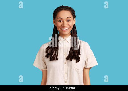 Porträt einer fröhlichen schönen Frau mit schwarzen Dreadlocks, mit charmanten einnehmenden Lächeln und positive Emotionen, trägt weißes Hemd. Innenaufnahme des Studios isoliert auf blauem Hintergrund. Stockfoto