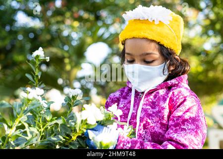 Mädchen Kind mit medizinischer Gesichtsmaske spielt mit Blumen im Park - Konzept der Gesundheitsversorgung, Umweltverschmutzung Sicherheitsmaßnahmen, Achtsamkeit und covid-19 Schutz. Stockfoto