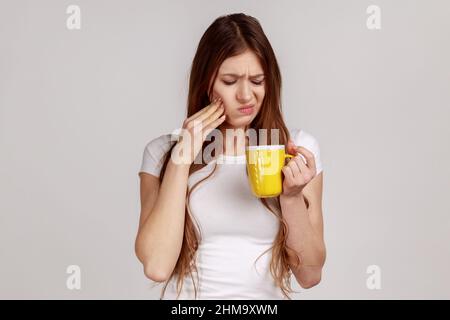 Kranke, ungesunde Frau, die das Kinn berührt, hat Zahnschmerzen, hat empfindliche Zähne, fühlt Schmerzen nach dem Trinken von kaltem Wasser, trägt ein weißes T-Shirt. Innenaufnahme des Studios isoliert auf grauem Hintergrund. Stockfoto