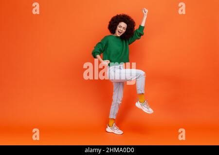 In voller Länge Porträt einer Frau mit Afro-Frisur trägt grünen lässigen Stil Pullover schreiend mit glücklichen Ausdruck, feiert Erfolg. Innenaufnahme des Studios isoliert auf orangefarbenem Hintergrund. Stockfoto