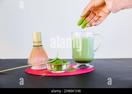 Crop anonyme weibliche Herstellung gesunder Matcha-Tee mit Milch in Glas Becher in der Nähe von Bambus Chasen und Chashaku auf dem Tisch vor weißem Hintergrund platziert Stockfoto