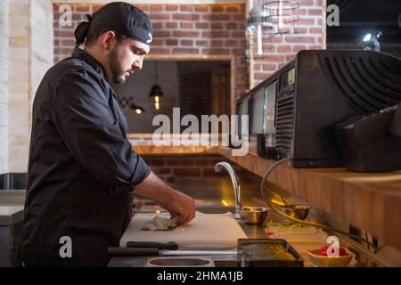 Seitenansicht eines konzentrierten bärtigen Chefkochs, der Reis auf Nori verteilt, während er während der Arbeit im Restaurant Sushi auf dem Schneidebrett zubereitet Stockfoto