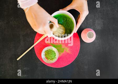 Draufsicht auf das anonyme Weibchen mit Bambus-Chasen, die Wasser schlagen und grünem Matcha-Pulver für ein gesundes japanisches Getränk auf einem hellen Kreis auf dem schwarzen Tisch Stockfoto