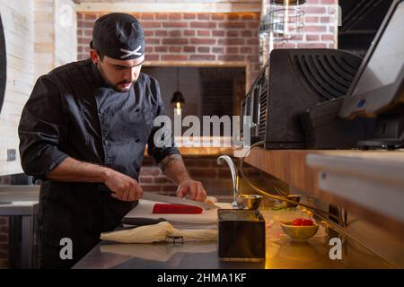 Konzentrierter männlicher Koch schneidet in Uniform rohen Fisch mit Messer, während er Sushi auf dem Tisch in der Nähe des Gebäckbeutels mit Frischkäse im Restaurant zubereitet Stockfoto