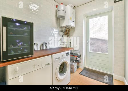 Weinkühlschrank mit Sammlung von alkoholischen Getränken in der Nähe der modernen weißen Waschmaschine in der Waschküche mit Boiler und Tür platziert Stockfoto