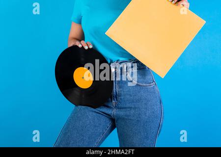 Beschnittene, nicht erkennbare weibliche Meloman mit Retro-Vinyl-Schallplatte und Papierverpackung, die auf blauem Hintergrund im hellen Studio steht Stockfoto