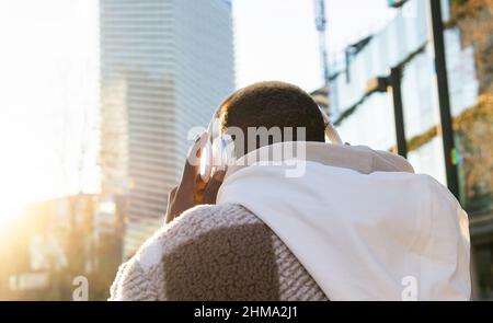 Rückansicht eines nicht erkennbaren Mannes mit kurzen Haaren, der Kopfhörer aufsetzt, während er Musik auf einer sonnigen Straße mit modernen Gebäuden hört Stockfoto