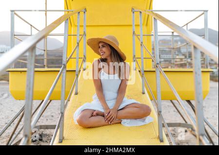 Entzückte Frau in Sommerkleid und Strohhut, die auf dem gelben Rettungsschwimmerturm am Sandstrand sitzt und wegschaut Stockfoto