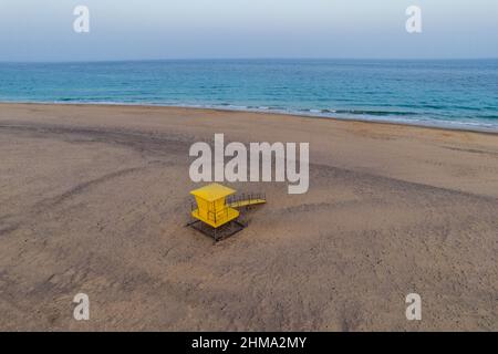 Spektakuläre Luftaufnahme des leuchtend gelben Rettungsschwimmerturms, der sich an einer sandigen Küste vor dem blauen Meer unter Sonnenuntergang befindet Stockfoto