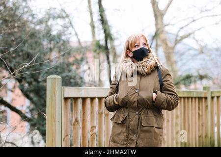 Frau in Oberbekleidung und schwarzer Schutzmaske, die während einer Pandemie am Herbsttag in der Stadt auf der Straße mit Bäumen und Holzzaun aufschaut Stockfoto
