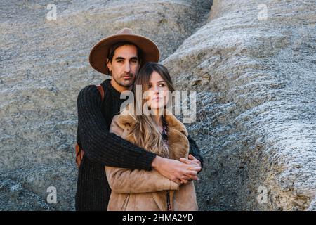 Hispanische Reisende in Hut umarmte Freundin, während sie bei Tageslicht in der Nähe von felsiger Oberfläche in der Natur zusammenstanden Stockfoto