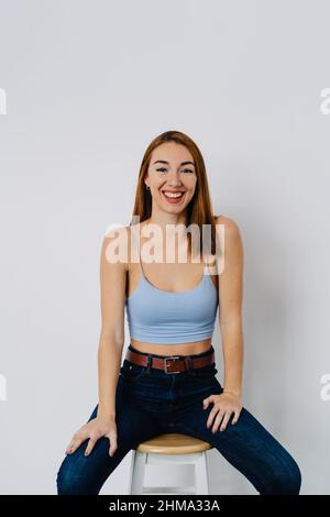 Positive junge Rothaarige, die Jeans und Oberteil trägt, sitzt auf einem Hocker nahe der weißen Wand und schaut auf die Kamera Stockfoto