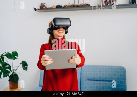 Lächelnde anonyme Frau in rotem Pullover, die die virtuelle Realität erkundet, während sie zu Hause ein Tablet nutzt und ein VR-Headset trägt Stockfoto
