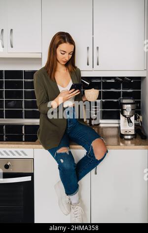 Fokussierte Frau mit roten Haaren in zerrissenen Jeans, die auf der Küchentheke mit einer Tasse Kaffee sitzt und das Smartphone durchstöbert Stockfoto