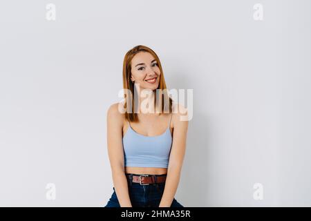 Positive junge Rothaarige, die Jeans und Oberteil trägt, sitzt auf einem Hocker nahe der weißen Wand und schaut auf die Kamera Stockfoto
