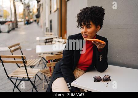 Moderne afroamerikanische Frau mit lockigen Haaren sitzt am Tisch im Café im Freien und zeichnet Sprachnachrichten auf dem Handy auf Stockfoto