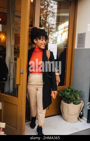 Positive afroamerikanische Frau in stilvollem Mantel mit Rucksack und Smartphone, das die Tür des Cafés öffnet Stockfoto