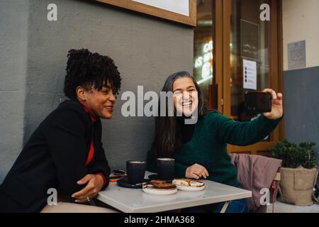 Positive ethnisch reife Frau mit Latte und Selbstporträt mit lachenden afroamerikanischen Freundin im Kaffeehaus im Freien Stockfoto