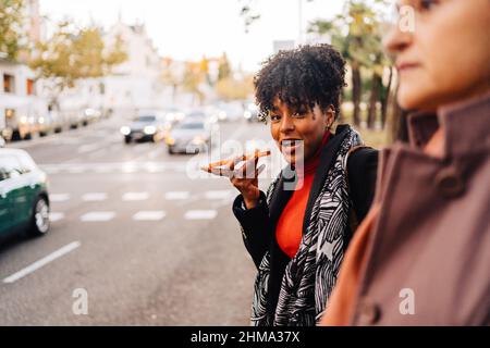 Inhalt afroamerikanische Frau, die Sprachnachrichten am Telefon aufzeichnet, während sie am Straßenrand der Stadt gegen verschwommene Frauen steht Stockfoto