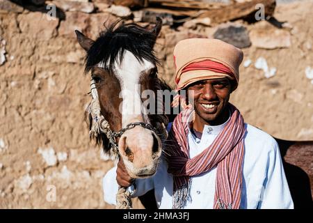 Fröhlicher arabischer Mann im Turban streichelt entzückendes Pferd, während er in der Nähe eines alten Steingebäudes in der Siedlung an einem sonnigen Tag steht Stockfoto