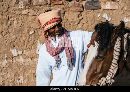 Fröhlicher arabischer Mann im Turban streichelt entzückendes Pferd, während er in der Nähe eines alten Steingebäudes in der Siedlung an einem sonnigen Tag steht Stockfoto